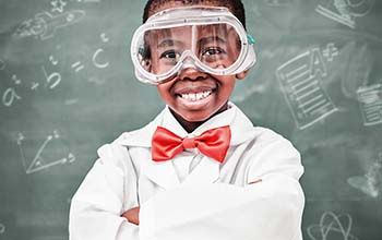 Boy wearing lab coat, red bow tie and safety goggles in a classroom.  Blackboard in the background with different science ico