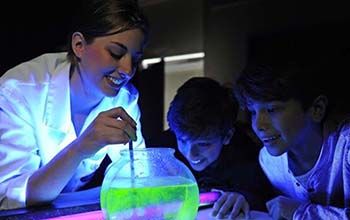 Mad Scientist pouring liquid that turns green with the use of a black light into a bowl of water with 2 kids looking on.